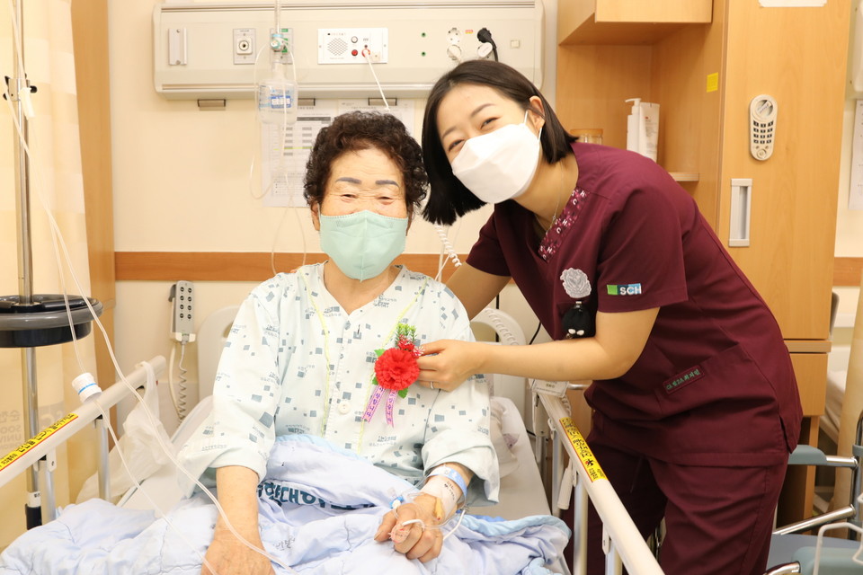 병동 간호사가 카네이션을 달아 드리고 어르신 환자와 함께 활짝 웃고 있다.